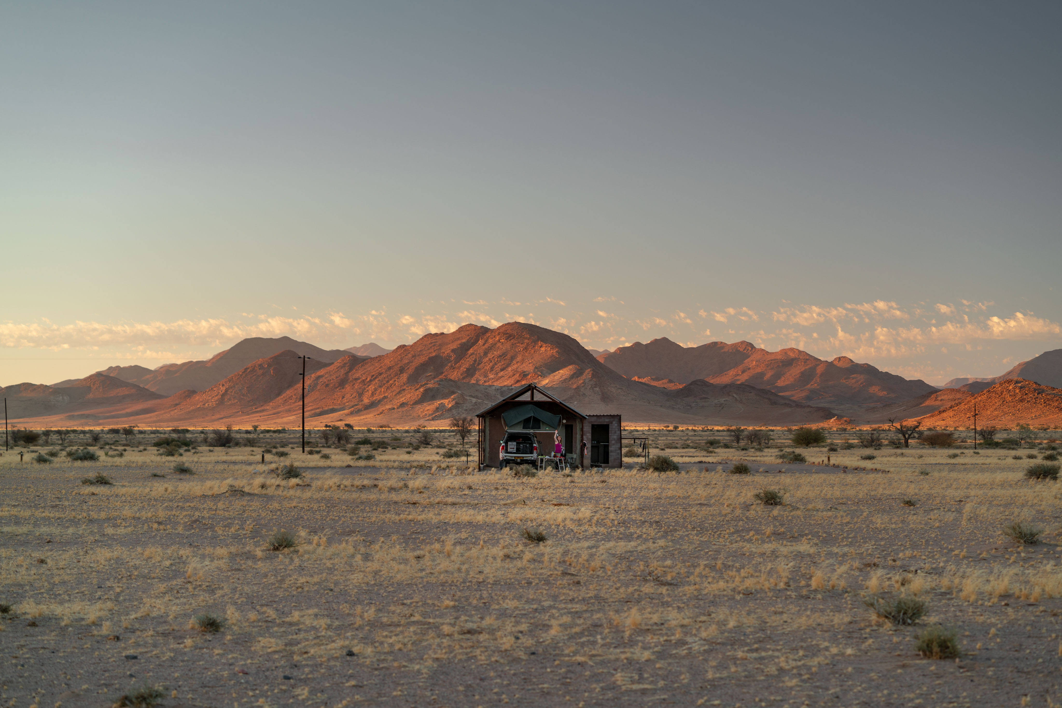 A wide desert plain at sunset. In the middle distance, a tiny petrol-station-style canopy with a single car parked beneath it and one figure standing beside. Behind, a long jagged ridge of mountains lit deep red-orange by the setting sun. Above, a band of soft pink cloud across a pale sky.