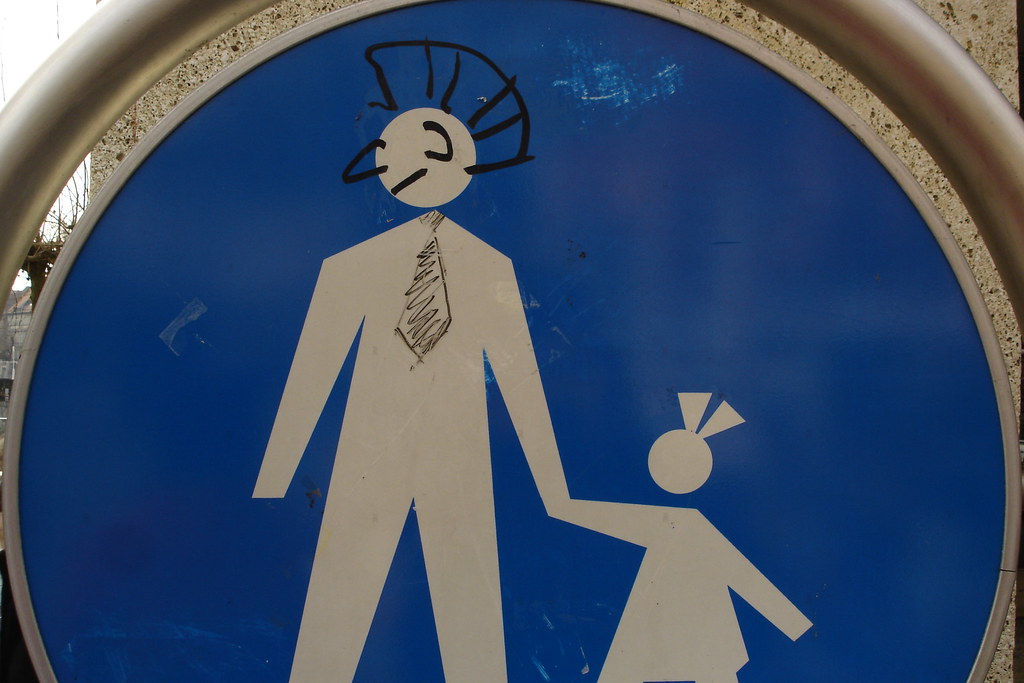 A blue circular pedestrian-crossing sign showing a white stick figure of a man holding the hand of a small child. Someone has drawn on it in black marker: a punk mohawk on the man's head, a sketched tie around his neck, and a small bow-clip in the girl's hair.