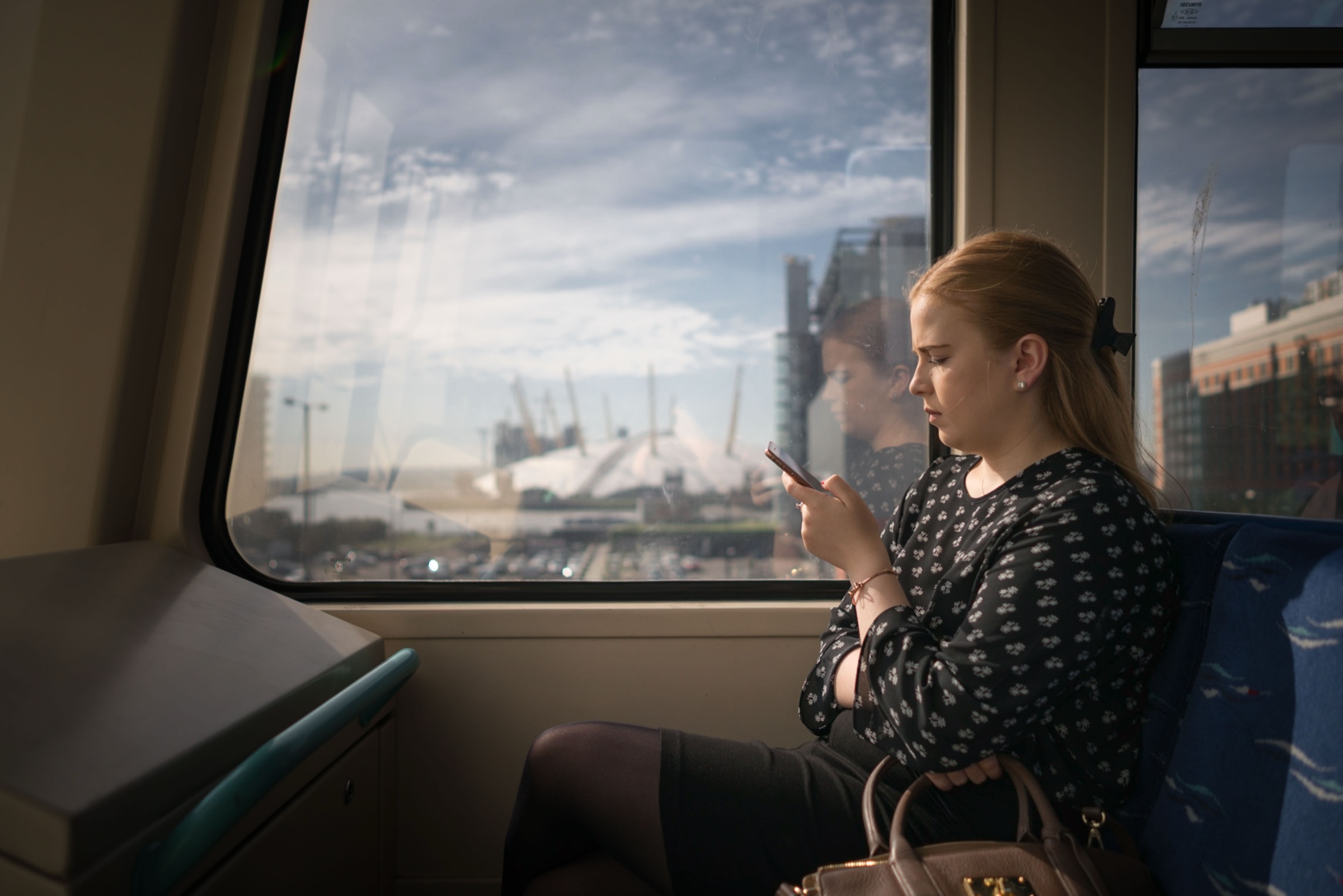 A young woman with red hair sits by the window of a London DLR train in late afternoon sun, looking down at her phone. Through the window behind her, the white domes of the O2 arena and the towers of Canary Wharf rise against a soft sky of broken cloud. Her own reflection floats faintly in the glass.