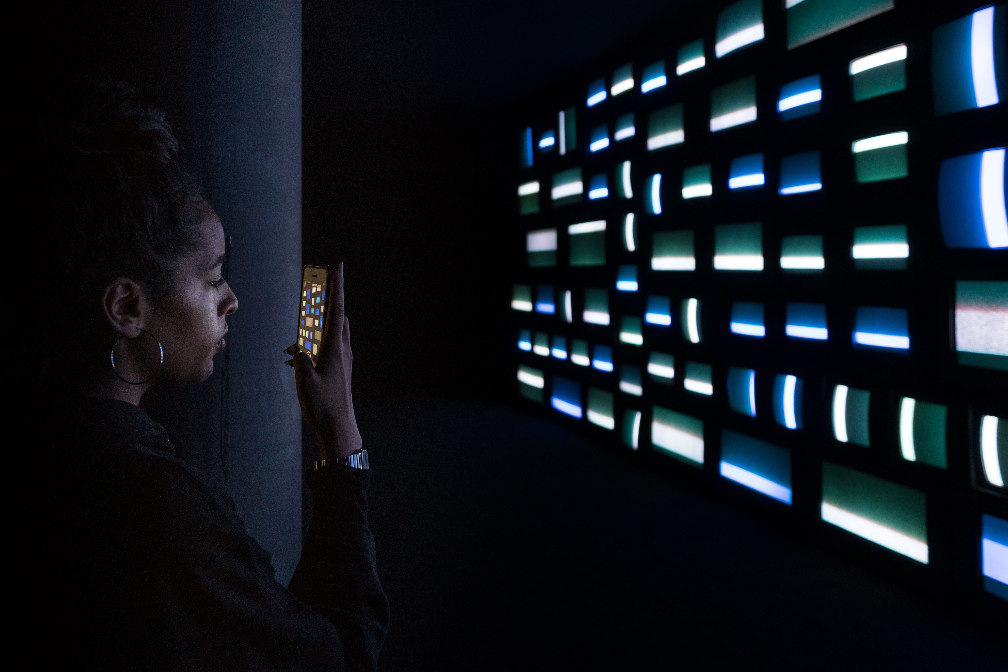 In a darkened gallery, a young woman with hoop earrings holds her phone up to photograph a wall of vintage cathode-ray television screens, all glowing in cool blue and green bands of test-pattern light. The phone's display reflects a small bright copy of the screens she is photographing.