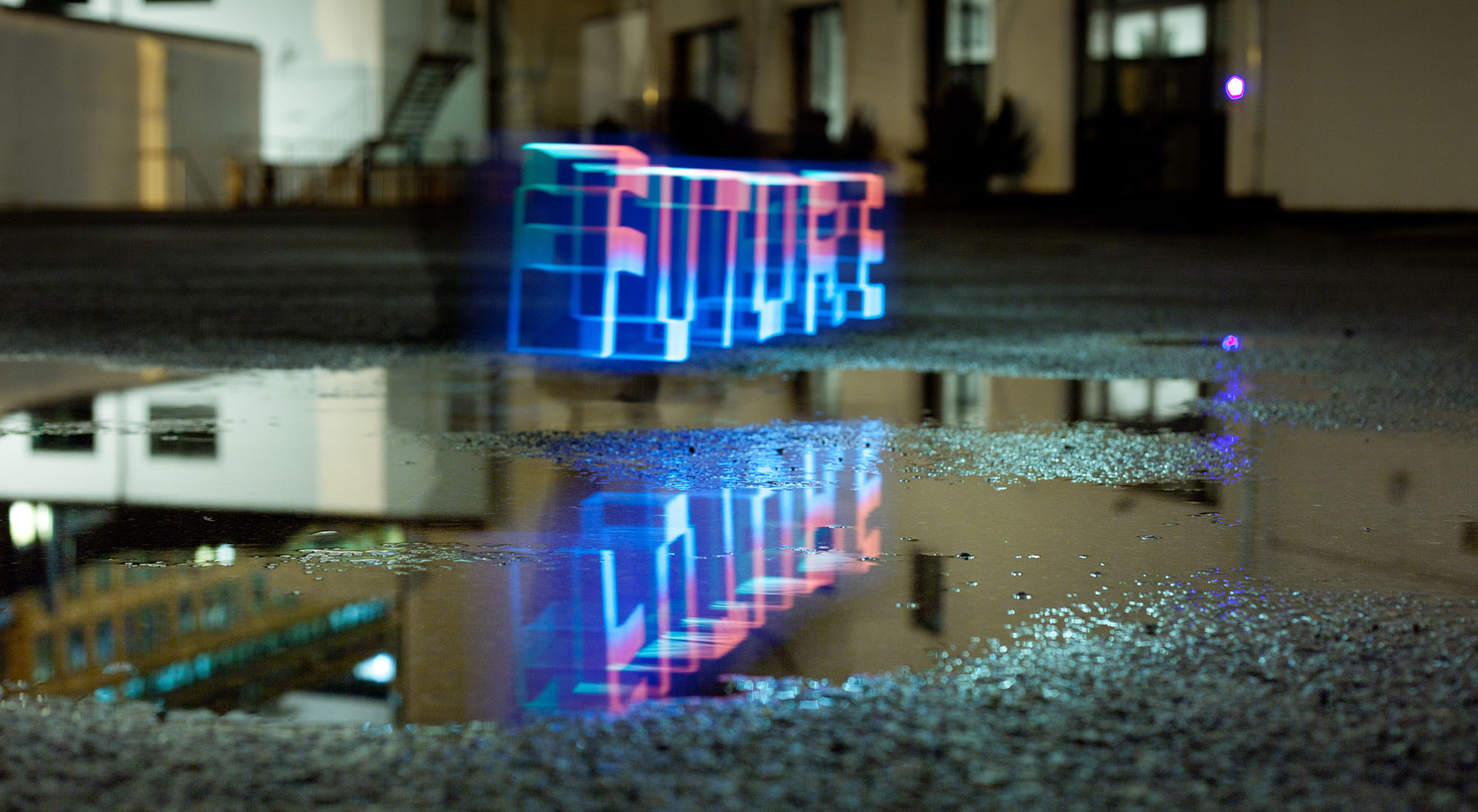 A long-exposure photograph at night: the word FUTURE drawn in 3D blue and pink light hovering above a wet courtyard, its perfect mirror image reflected in a puddle on the asphalt below. The surrounding industrial building, doors, and a single magenta lamp are reflected alongside it.
