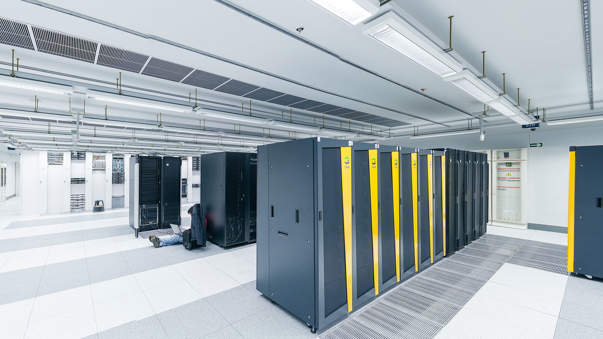 Interior of the data centre: a wide aisle flanked by tall black server racks, lit in cold blue