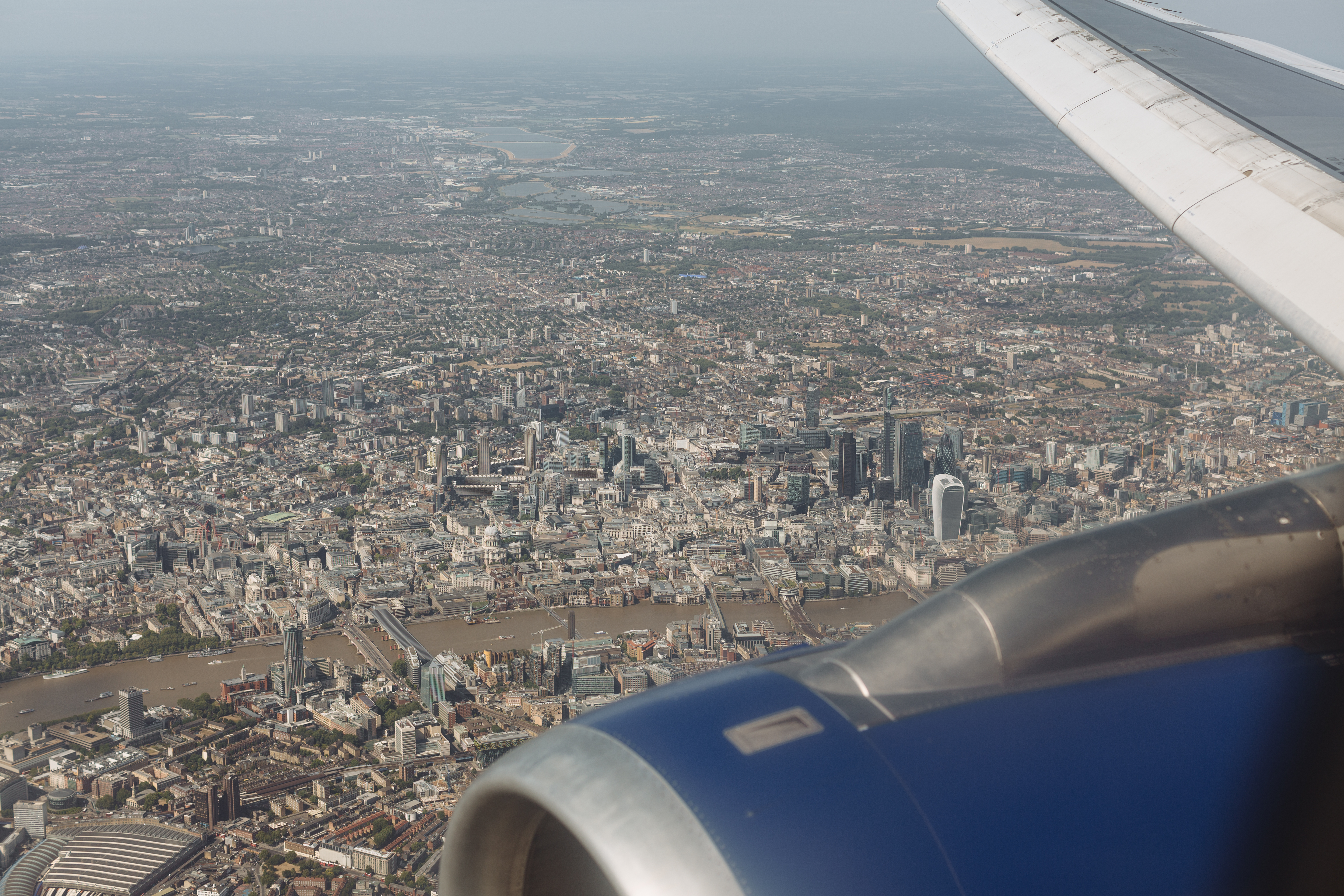An aerial photograph of central London on a hazy July afternoon, taken through the window glass of an aircraft circling in a holding pattern. The Thames meanders through the lower half of the frame; the City of London cluster — the Shard, the Walkie-Talkie, the Cheesegrater, the Gherkin — sits in the middle distance with Canary Wharf in the upper right; north London terraces stretch into haze along the upper edge.