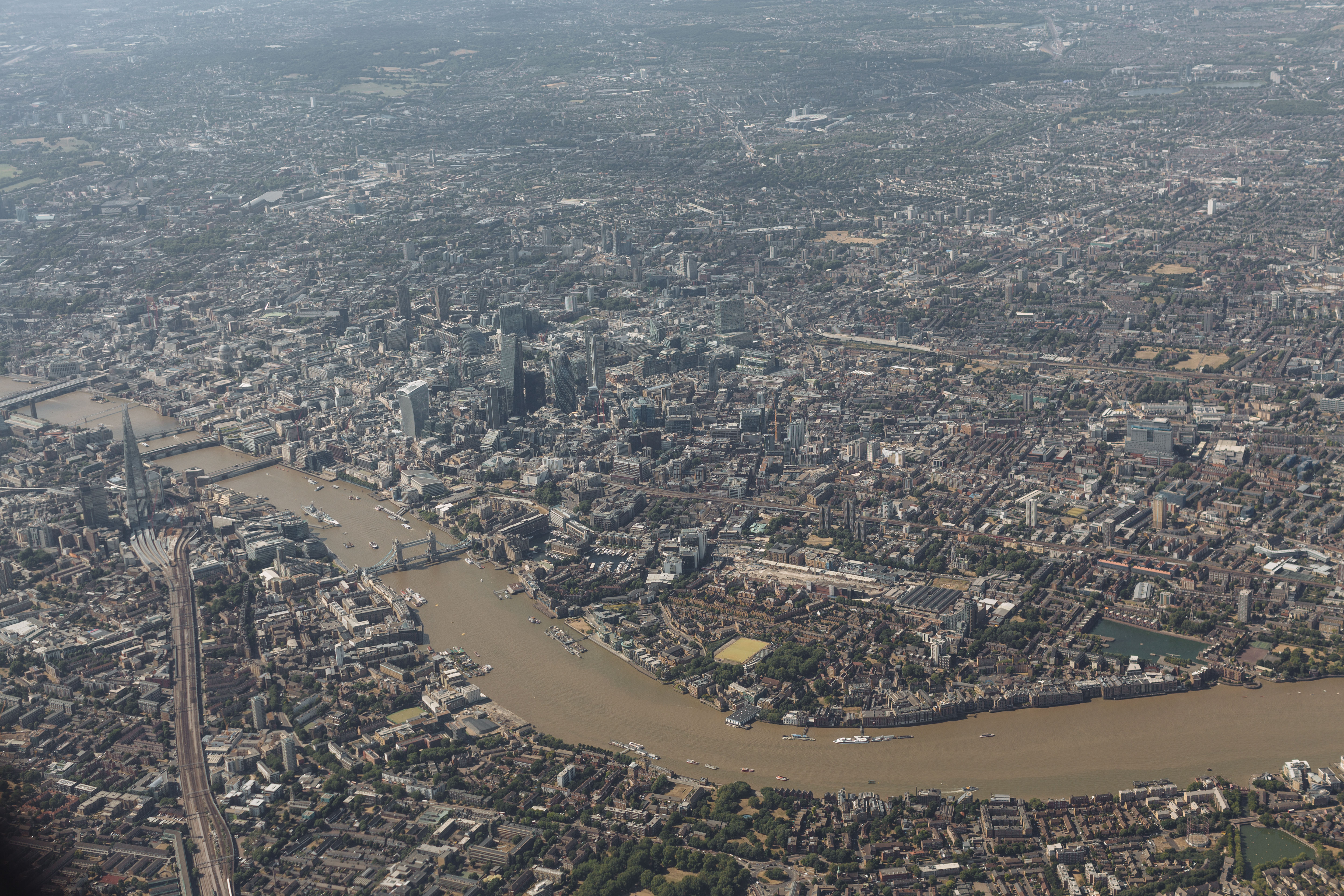 An aerial of east London during a banking turn. The Thames bends through the lower-right of the frame, Canary Wharf catches afternoon light at centre, the Greenwich peninsula and the O2 dome are tucked behind in the upper right. The horizon line is tilted slightly because the plane is mid-bank.