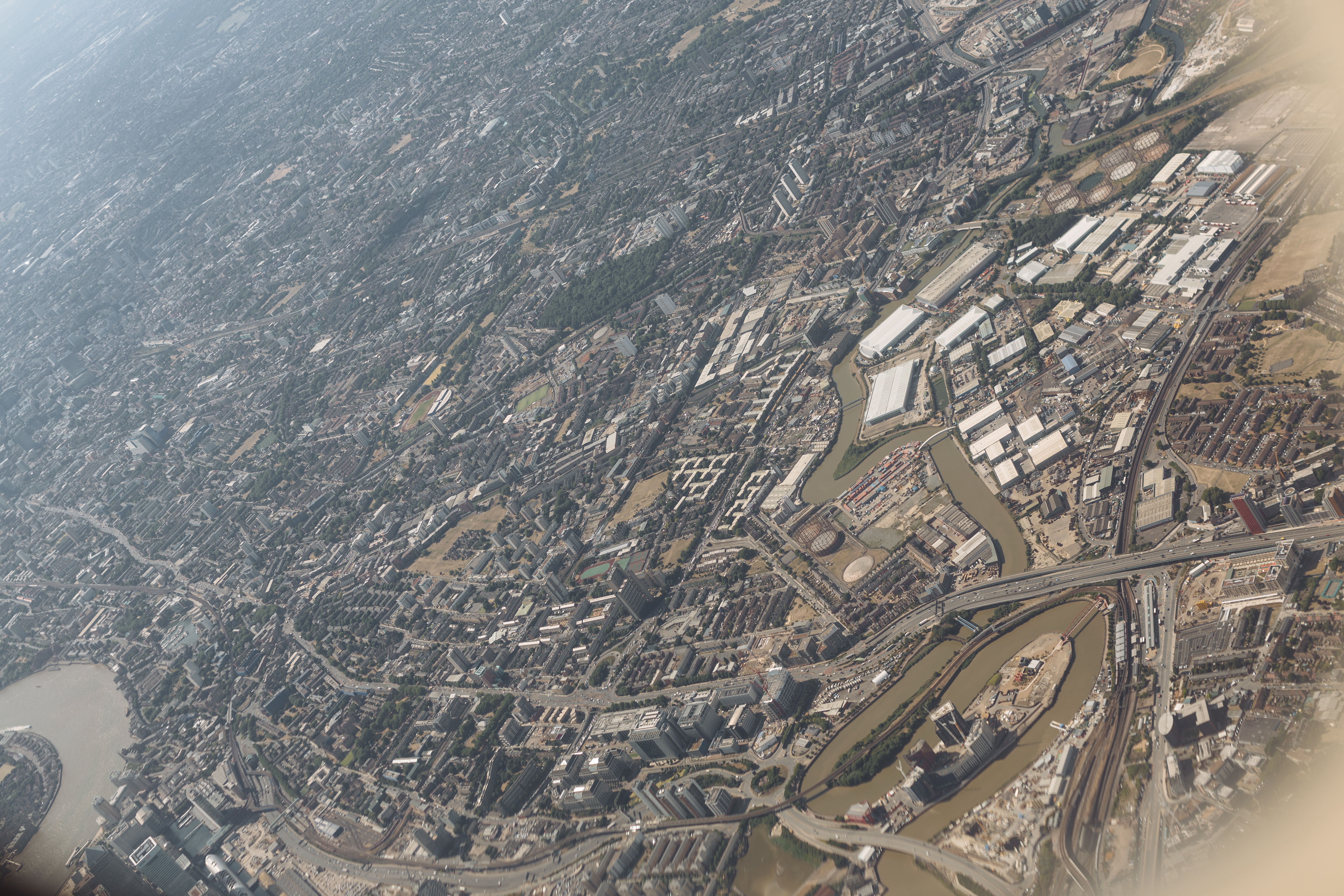 A near-vertical aerial of the Isle of Dogs and Canary Wharf. The Thames forms a perfect U around the peninsula, container ships visible in the West India Docks, the cluster of towers at the centre of the U lit by mid-afternoon sun. A faint reflection of the cabin window catches the upper edge of the frame.