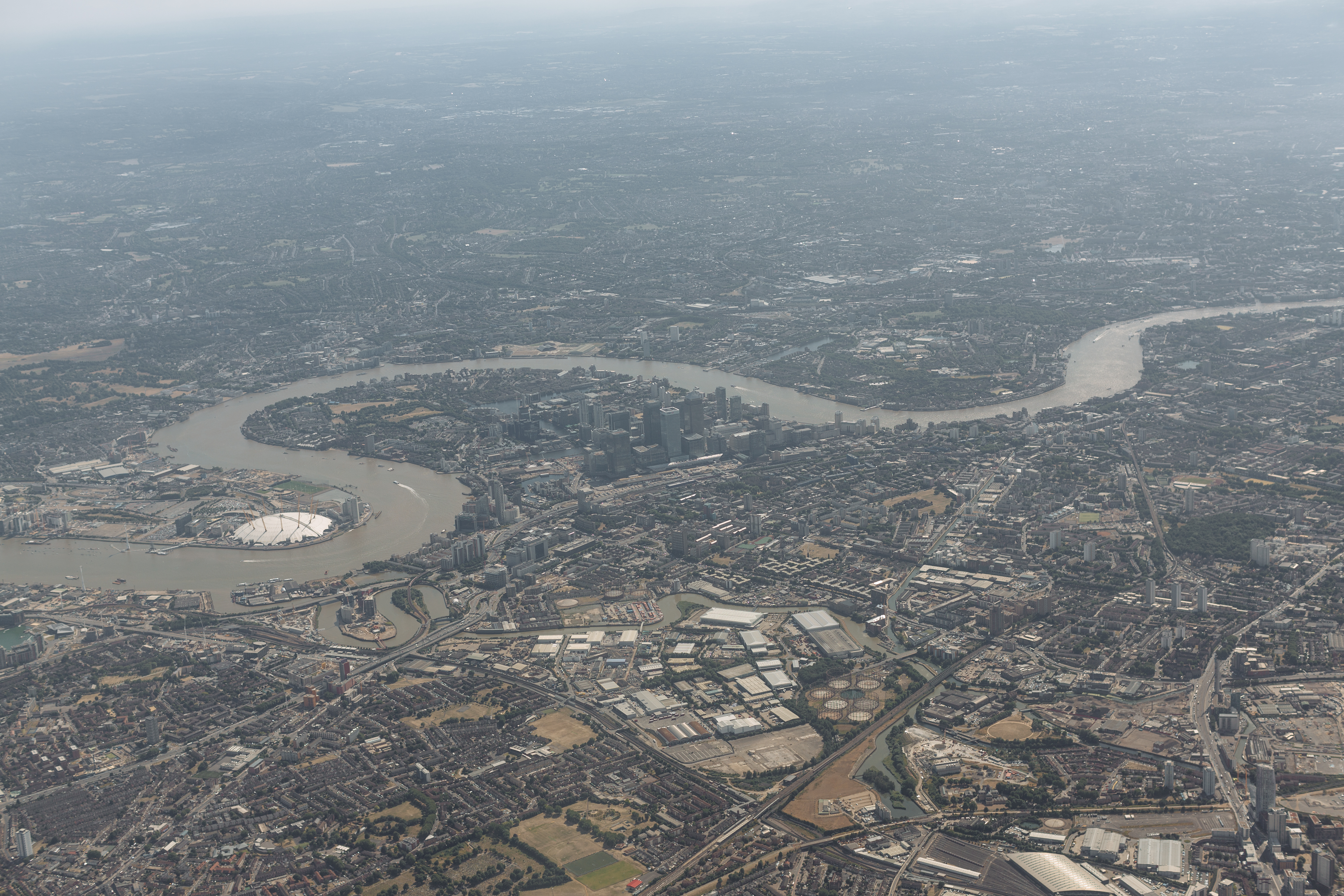 An aerial of central London catching late-afternoon sun on the City cluster. The Shard rises sharply at left of centre, Tower Bridge crosses the Thames in the foreground, the Walkie-Talkie and the Gherkin sit in the middle distance, the City stretches into hazy distance toward the upper edge. South Bank and Tate Modern at lower-left bank of the river.