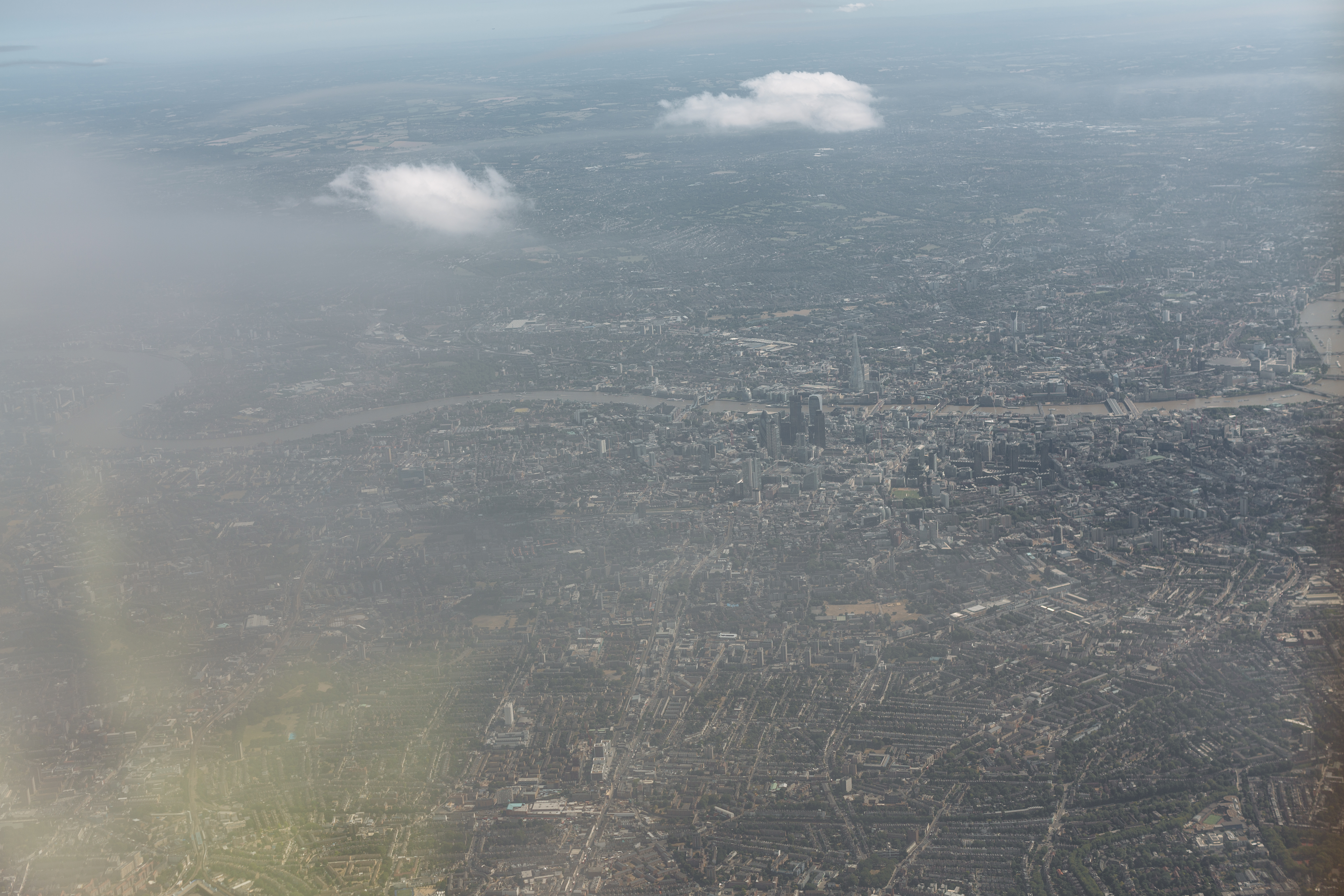 The final source frame: central London at hazy distance, the Thames running diagonally across the upper half, the City cluster at upper right. The aircraft wing and engine cowling intrude from the bottom-right corner of the frame as the plane completes the second bank.