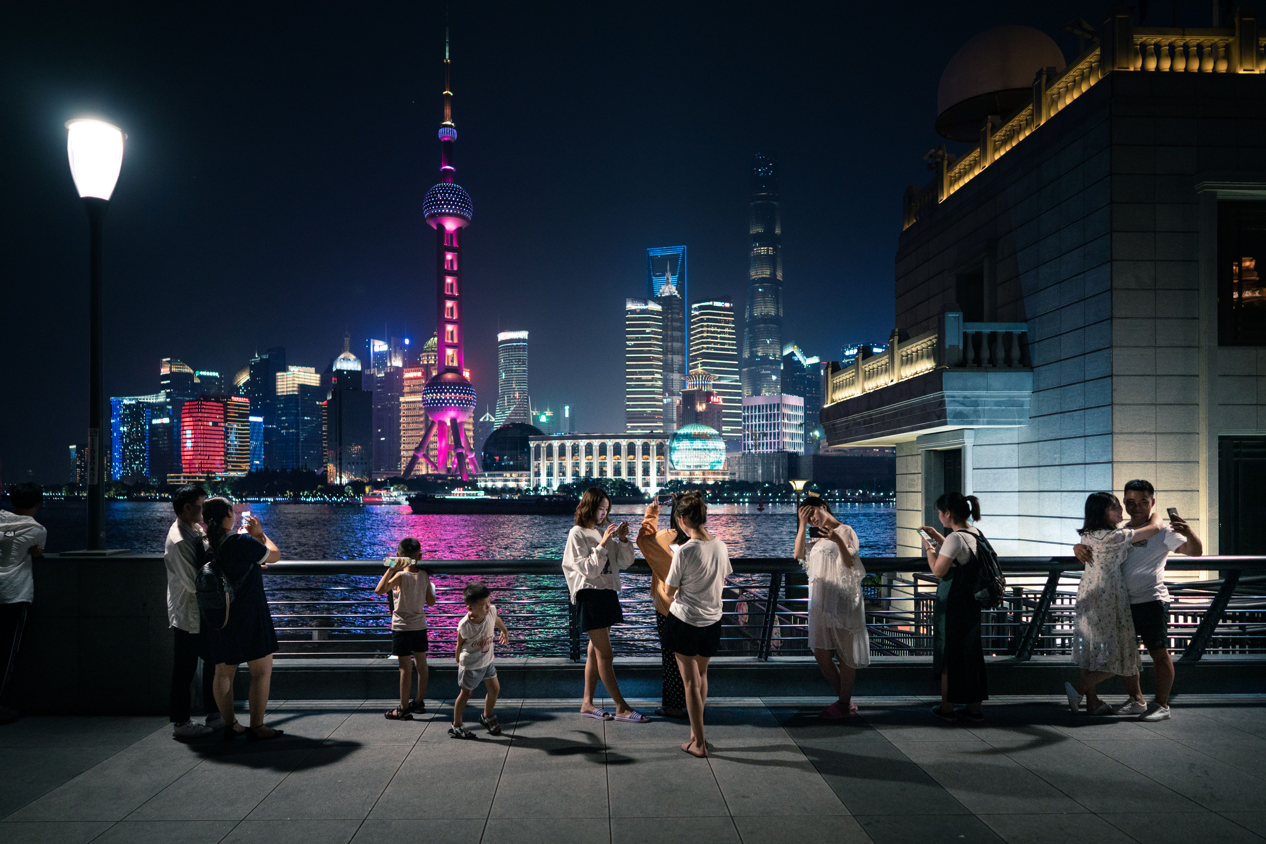 A line of people stand against a railing on the Shanghai Bund at night, every one of them holding up a phone. Across the dark Huangpu river, the Oriental Pearl Tower glows hot pink and the towers of Pudong are lit in saturated colour. A street lamp at left, a stone-clad guardhouse at right, the pavement empty in the foreground except for one running child.