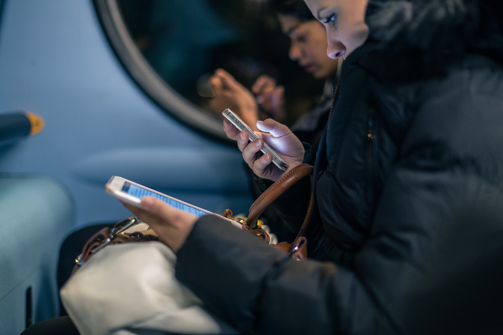 A close, shallow-depth-of-field photograph of two strangers on a metro train. A woman in a black puffer coat holds a white iPad in one hand and a small phone in the other, both screens lit. Across from her, partly out of focus, a young man in a coat looks down at his own phone.