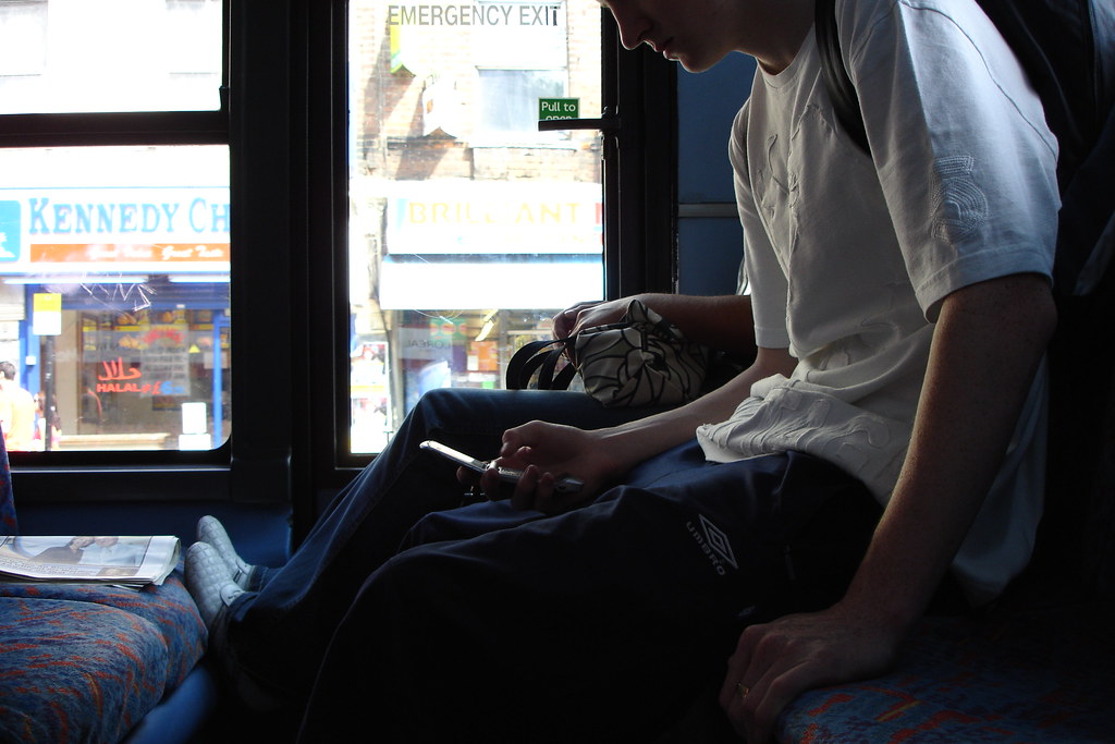 A young man in an Umbro tracksuit and a white t-shirt sits on a Manchester bus in afternoon light, head down, both hands cradling a small flip phone in his lap. Through the window beyond him: a Kennedy Chicken shopfront, a Halal sign, a takeaway whose lit yellow letters read 'BRILLIANT'. A newspaper lies face-down on the seat beside him.