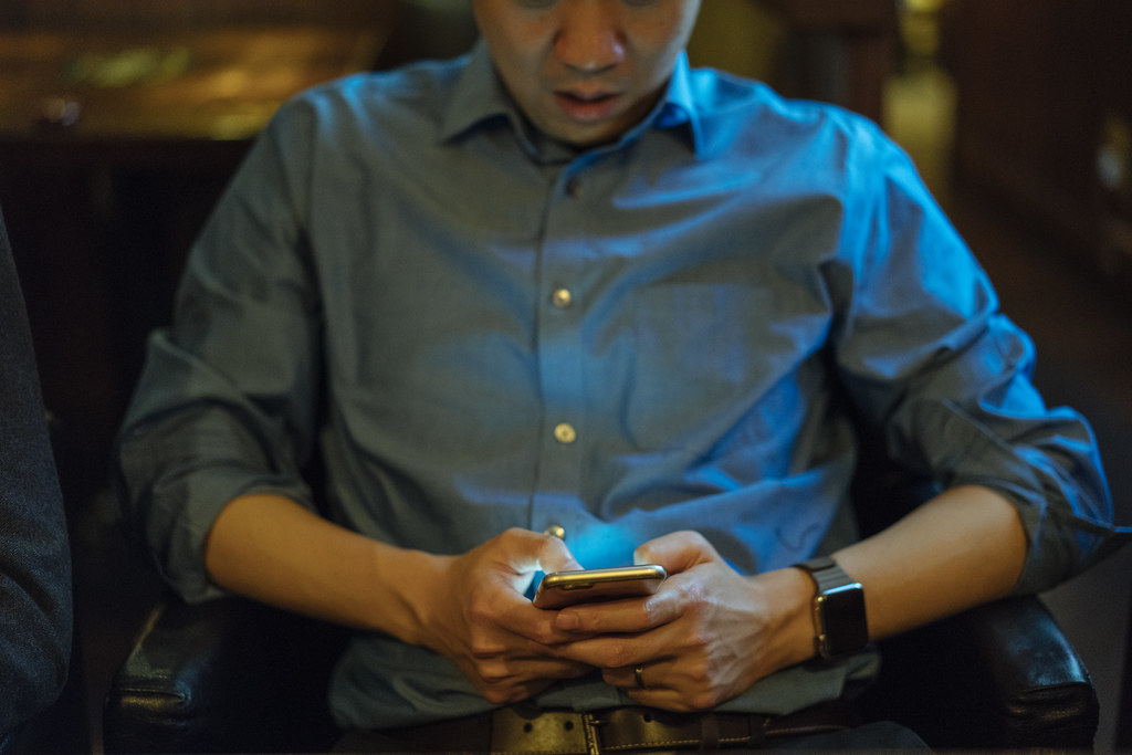 A close mid-body portrait of a man in a blue chambray shirt sitting in a leather club chair in soft warm hotel-lobby light. He cradles an iPhone in both hands, looking down at the screen. An Apple Watch is fastened on his right wrist; the screen casts a small cool blue glow up onto his forearms.