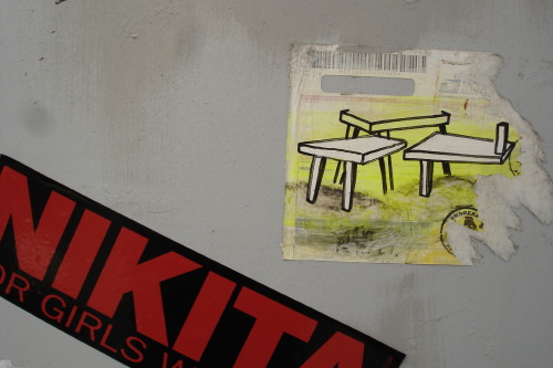A torn paste-up on a pale grey wall in Reykjavik: a small painting of two awkward stool-like wooden figures on a yellow ground, the paper around it scratched and peeling. Beside it, a slice of a red NIKITA sticker with the partial text 'OR GIRLS W', the streetwear brand sharing the surface with the hand-painted piece. One of seven frames made across the city in a single day.