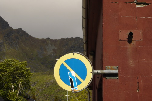 A circular Norwegian no-stopping sign (yellow rim, blue ground, red diagonal slash) bracketed off the wall of a deep red-painted building in Svolvær. The sign carries a small white-and-blue stickered figure pasted across its face. Behind, the dark mountains of the Lofoten archipelago and a heavy grey sky.