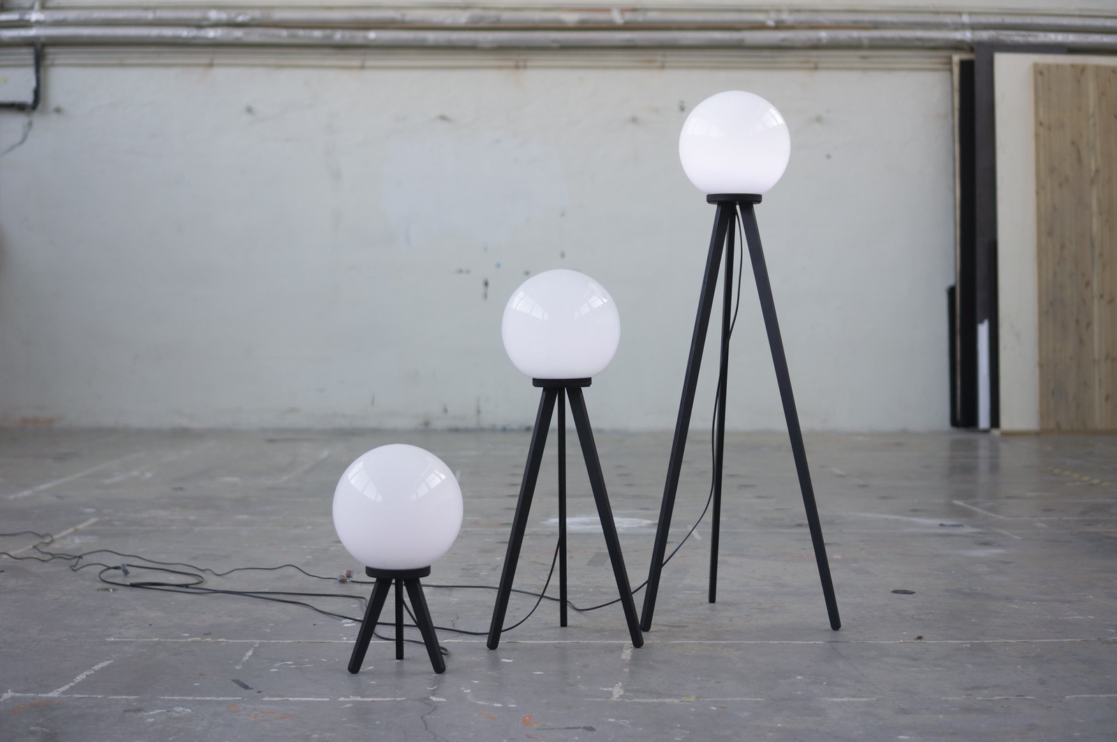 A cluster of Satellite Lamps in a gallery space, each a small tripod-mounted lamp glowing at different brightnesses as it samples GPS signal strength in its location