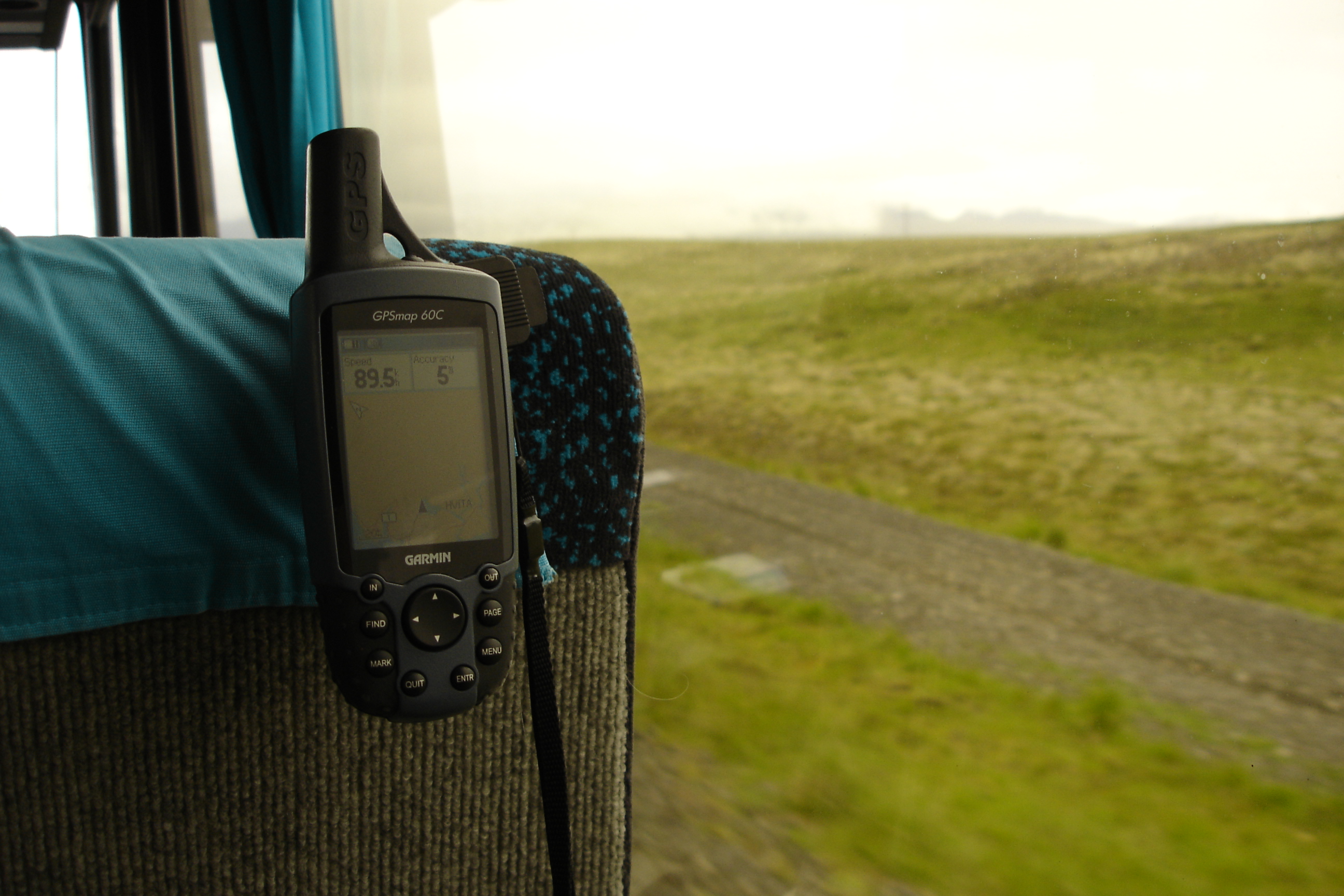 A Garmin GPSmap 60C clipped by its lanyard to the back of a bus seat. Screen reads 89.5 km/h. Through the window: empty Icelandic landscape, low road, distant hills in haze.