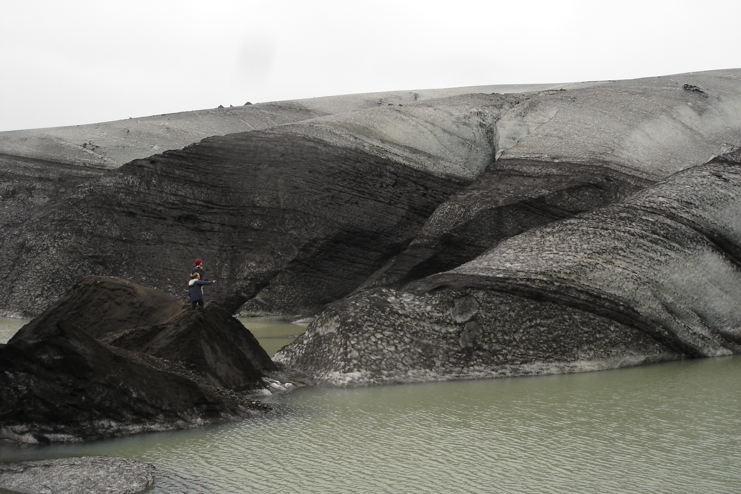 Two figures, a person carrying a child on their shoulders, on the dark moraine of a glacier. Stripes of black ash and grey ice; a pool of pale green meltwater.