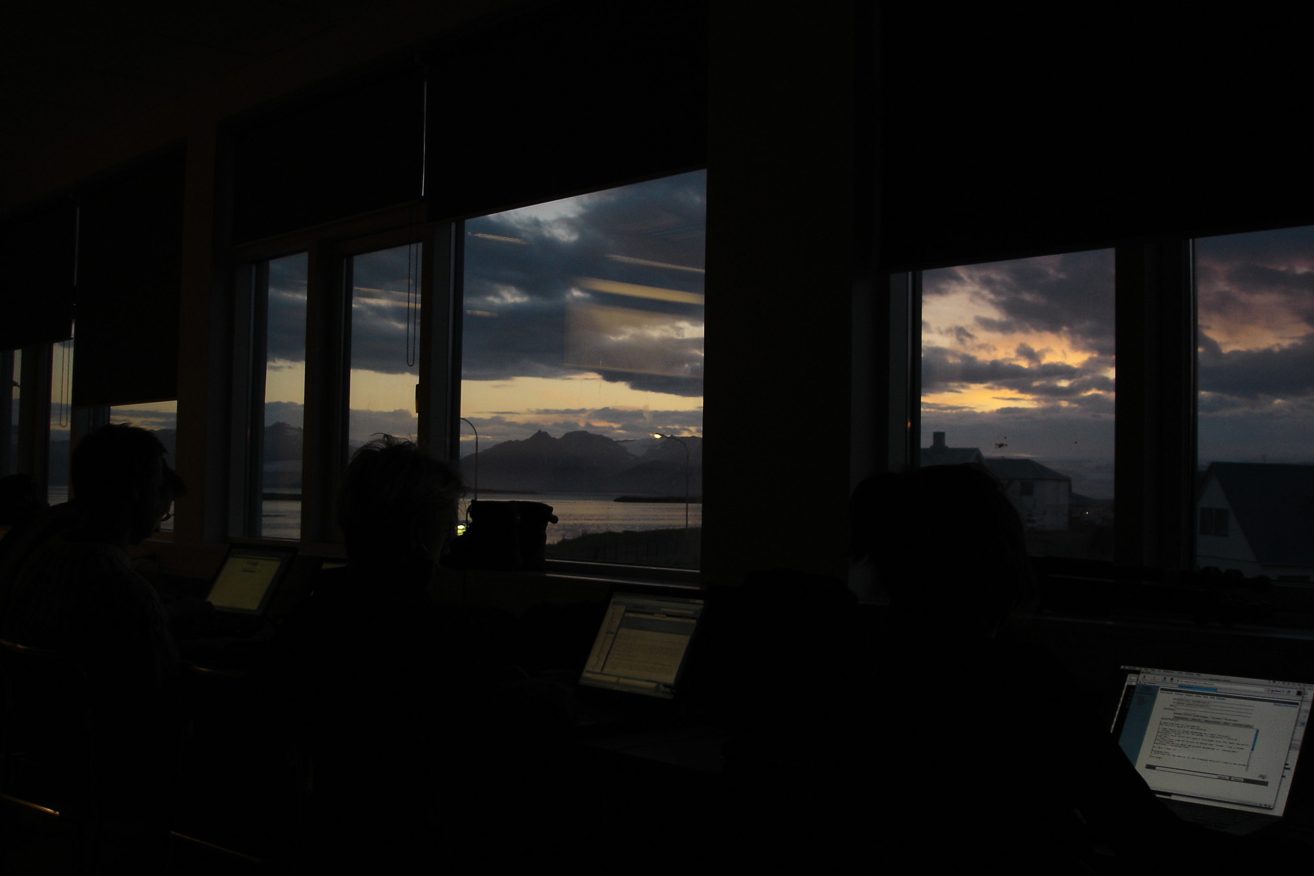 Silhouettes of three workshop participants at their laptops, lit only by their screens. Through the wall of windows behind: a fjord at the long subarctic dusk.