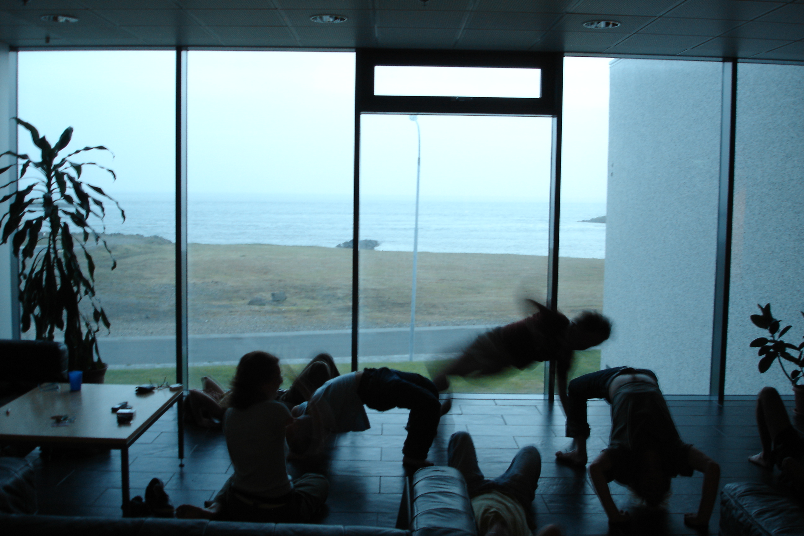 Workshop participants improvising breakdancing in the common room — one captured mid-air, motion-blurred. Beyond the floor-to-ceiling glass: low Icelandic coastline, the sea.