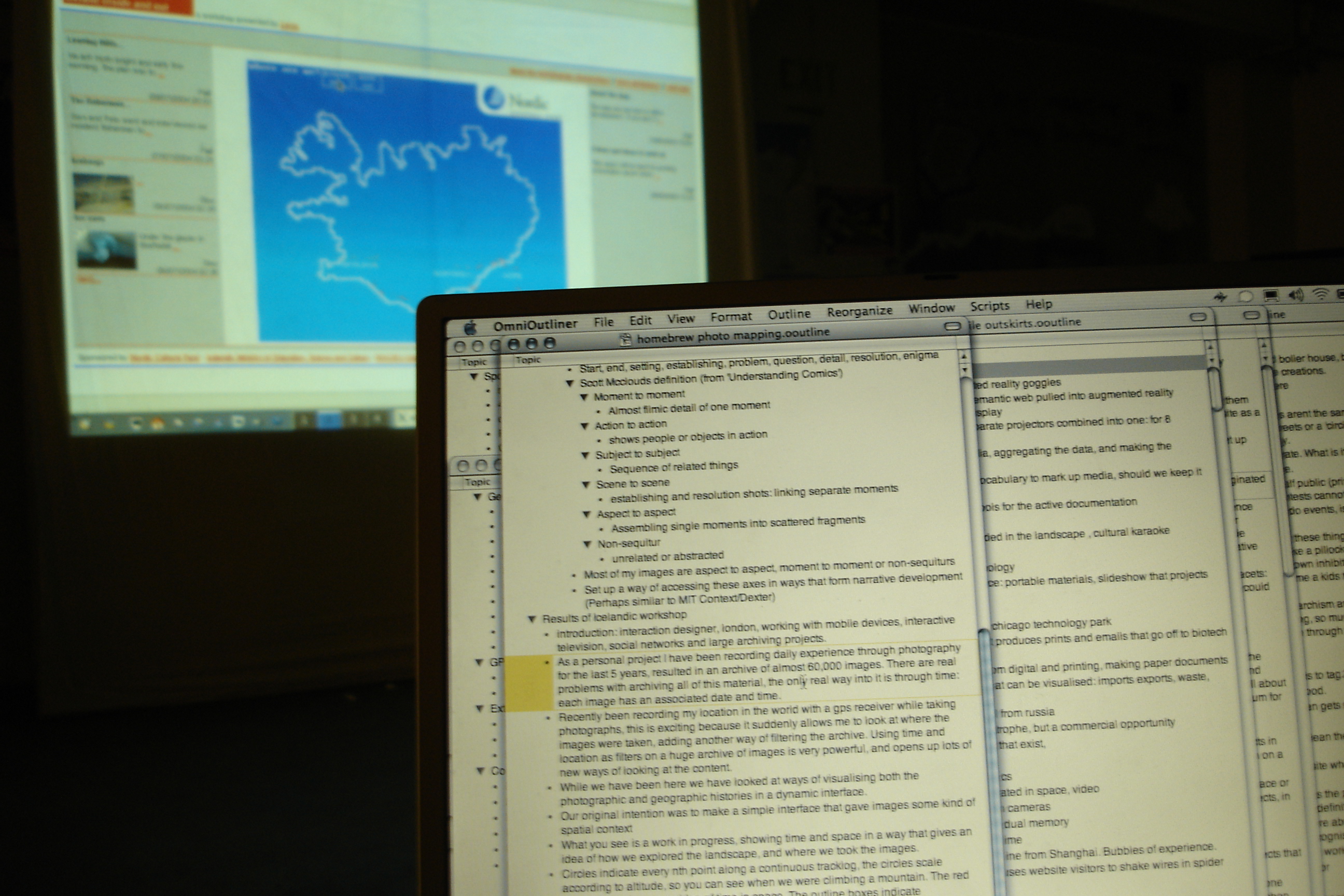 A laptop screen showing an OmniOutliner document with hand-typed notes about photo-mapping. Projected on the wall behind: a web page showing the white outline of Iceland with the GPS track of the trip overlaid as a continuous line.